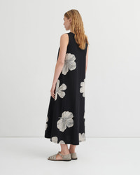 A woman stands with her back to the camera wearing a sleeveless black midi dress with a large white floral print, paired with gray strappy flat sandals, against a plain white studio background.