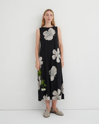 A woman stands against a plain white studio wall wearing a sleeveless black midi dress with a large white floral print. She is holding a small green sprig and wearing flat tan sandals.
