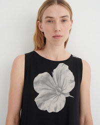 A woman wearing a sleeveless black top with a large gray watercolor-style hibiscus flower graphic on the front, photographed against a plain light gray background.