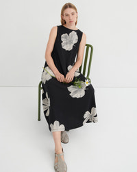 A woman sits on a green metal chair wearing a sleeveless black midi dress with a large white floral print. She is holding a small white flower and wearing gray strappy sandals against a clean white studio background.