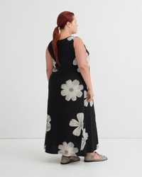 A woman with red hair tied back in a ponytail is shown from the back wearing a sleeveless black maxi dress with a large white floral pattern, paired with gray sandals, against a plain white studio background.
