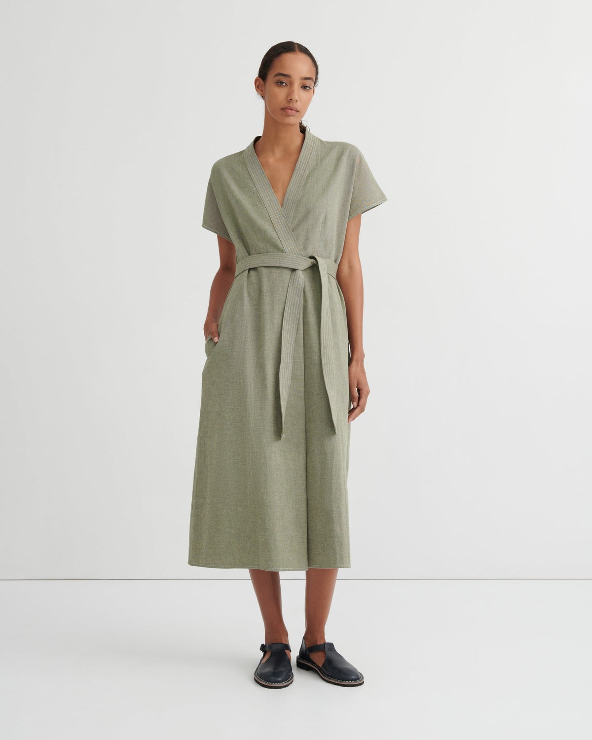 A woman modeling a sage green short-sleeve wrap dress with a matching waist tie, standing against a plain white studio background.