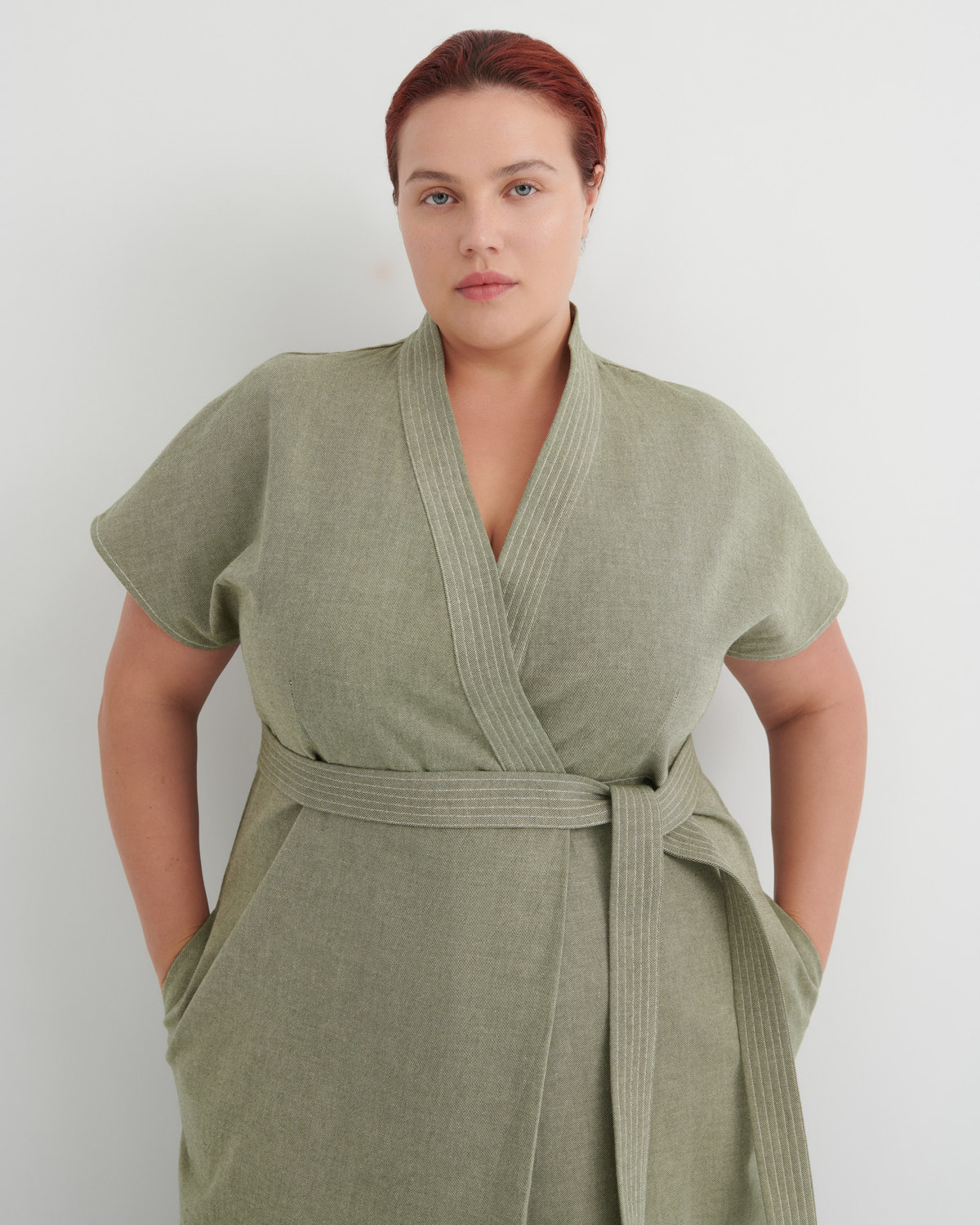 A woman wearing a sage green short-sleeve wrap robe with a matching waist tie, standing against a plain light gray background.