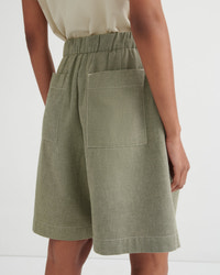 A close-up back view of a model wearing sage green linen shorts with an elastic waistband and large patch pockets, paired with a light beige top against a white studio background.