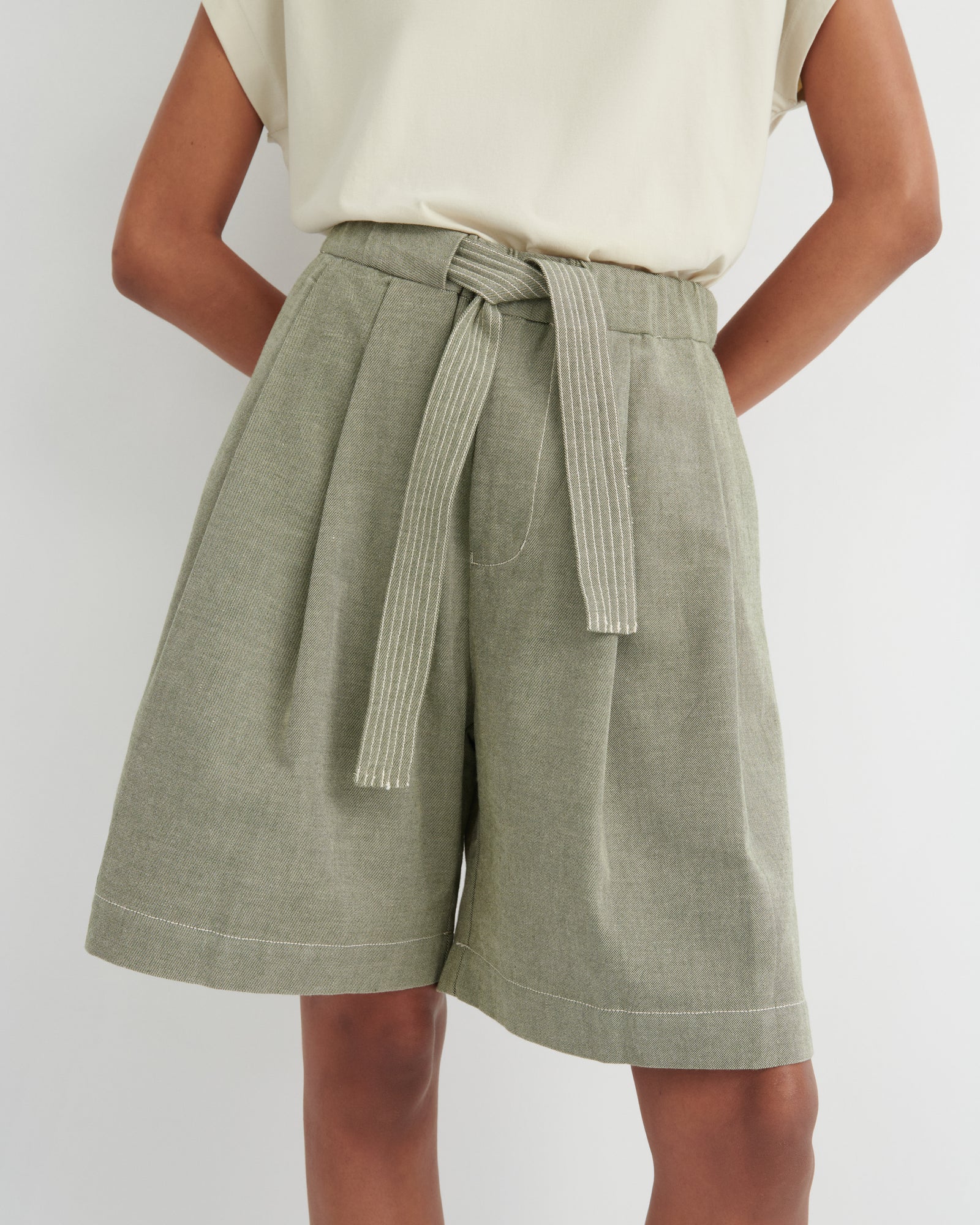 A person wearing high-waisted olive green pleated shorts with a matching tie belt, paired with a cream sleeveless top, against a light gray studio background.