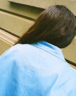 A person viewed from behind wearing a light blue Kowtow overshirt, with their dark brown hair visible against a beige wall background.