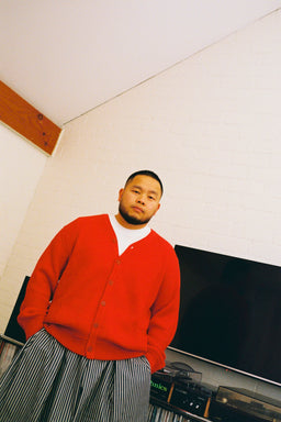 A man wearing a bright red knit cardigan over a white t-shirt and gray and white vertically striped pants, standing in a room with a white brick wall and a television in the background.