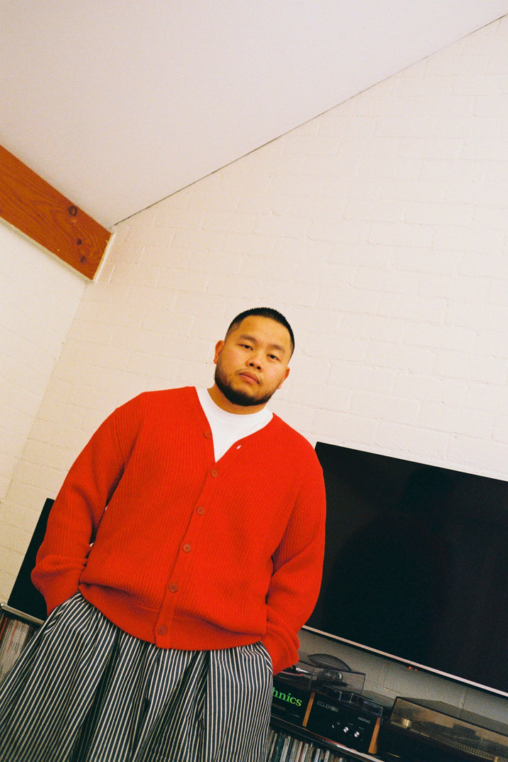 A man wearing a bright red knit cardigan over a white t-shirt and gray and white vertically striped pants, standing in a room with a white brick wall and a television in the background.