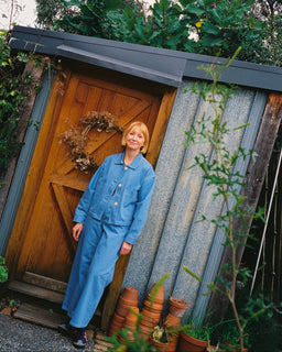 A person wearing a matching blue denim jacket and wide-leg pants set, standing outdoors next to a wooden door and stacked terracotta pots.