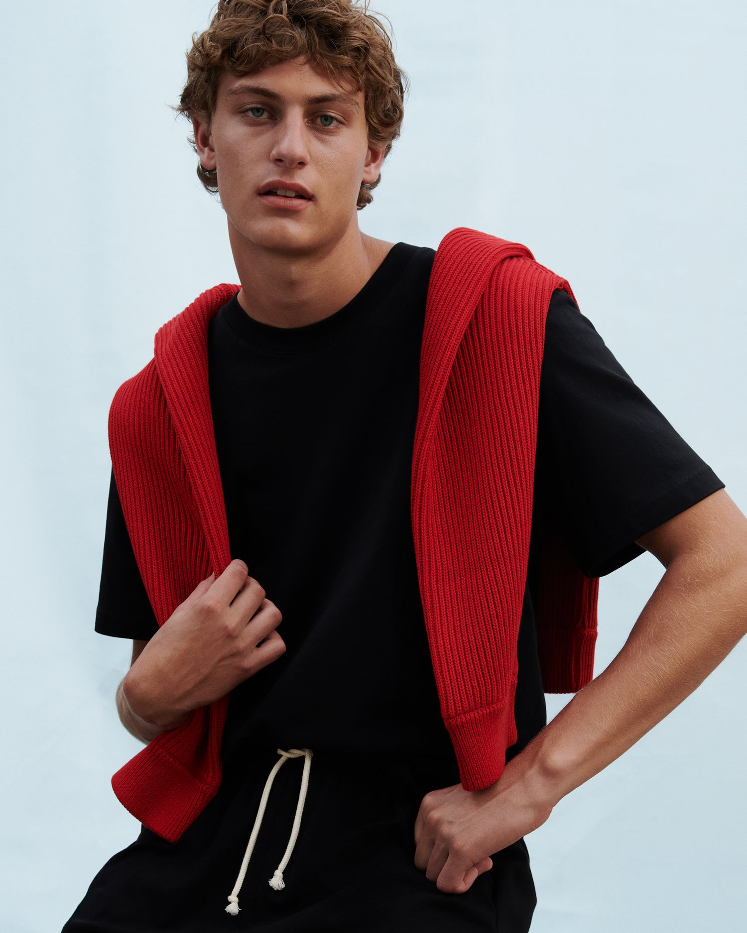 Model with blue background wearing a relaxed black t-shirt with a red sweater draped over his shoulders.