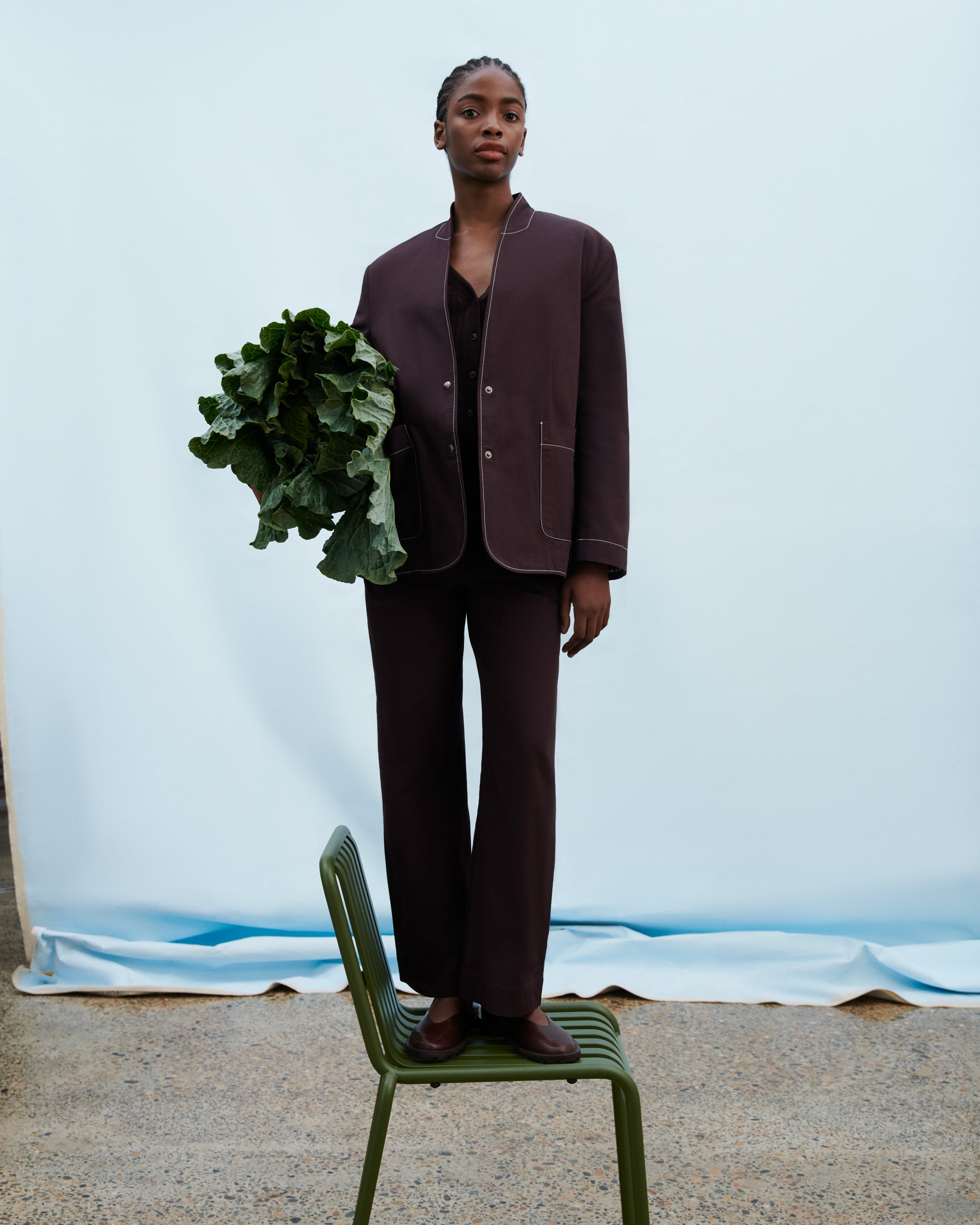 A model stands on a green metal chair wearing a dark brown two-piece suit with a cropped jacket and wide-leg trousers, holding a bunch of green leafy vegetables against a light blue studio backdrop.