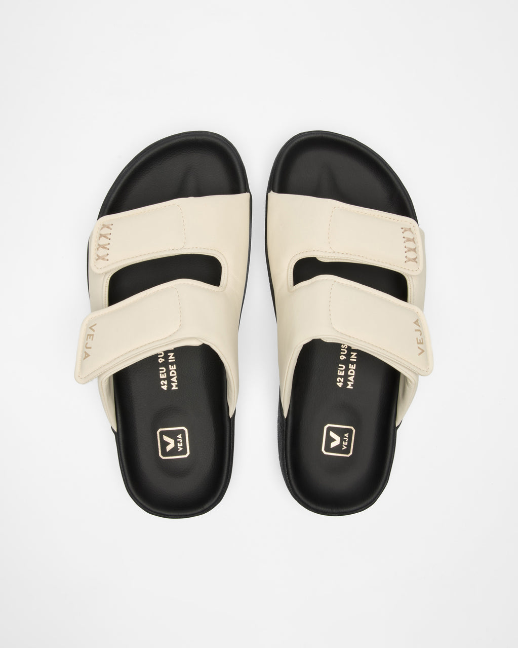 A pair of slide sandals with cream-colored straps and black footbeds, shown from a top-down view on a light gray background.