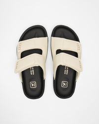 A pair of slide sandals with cream-colored straps and black footbeds, shown from a top-down view on a light gray background.