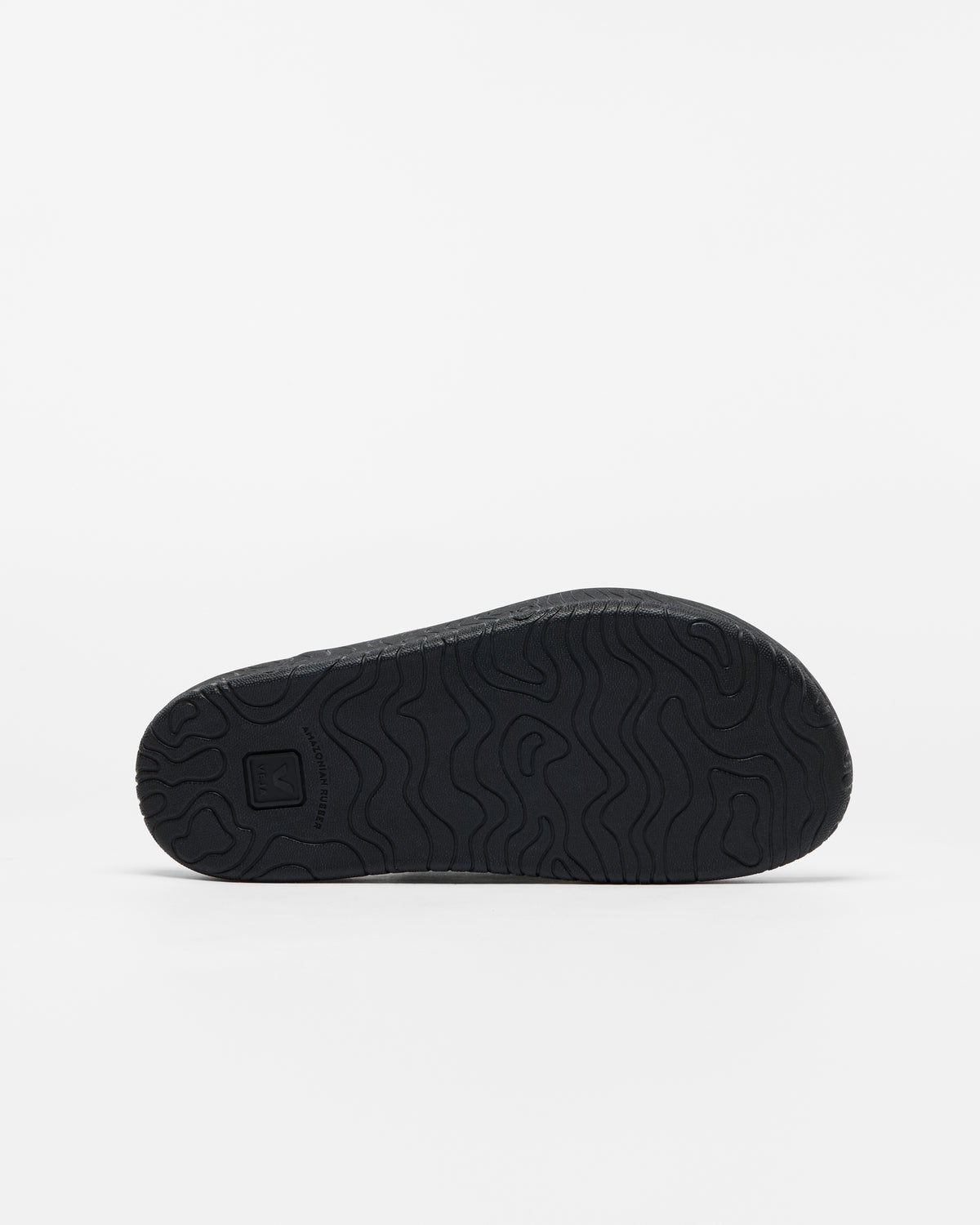 A close-up, bottom view of a black slip-on shoe's textured outsole, showing a wavy tread pattern and a small square logo embossed on the heel, set against a plain white background.