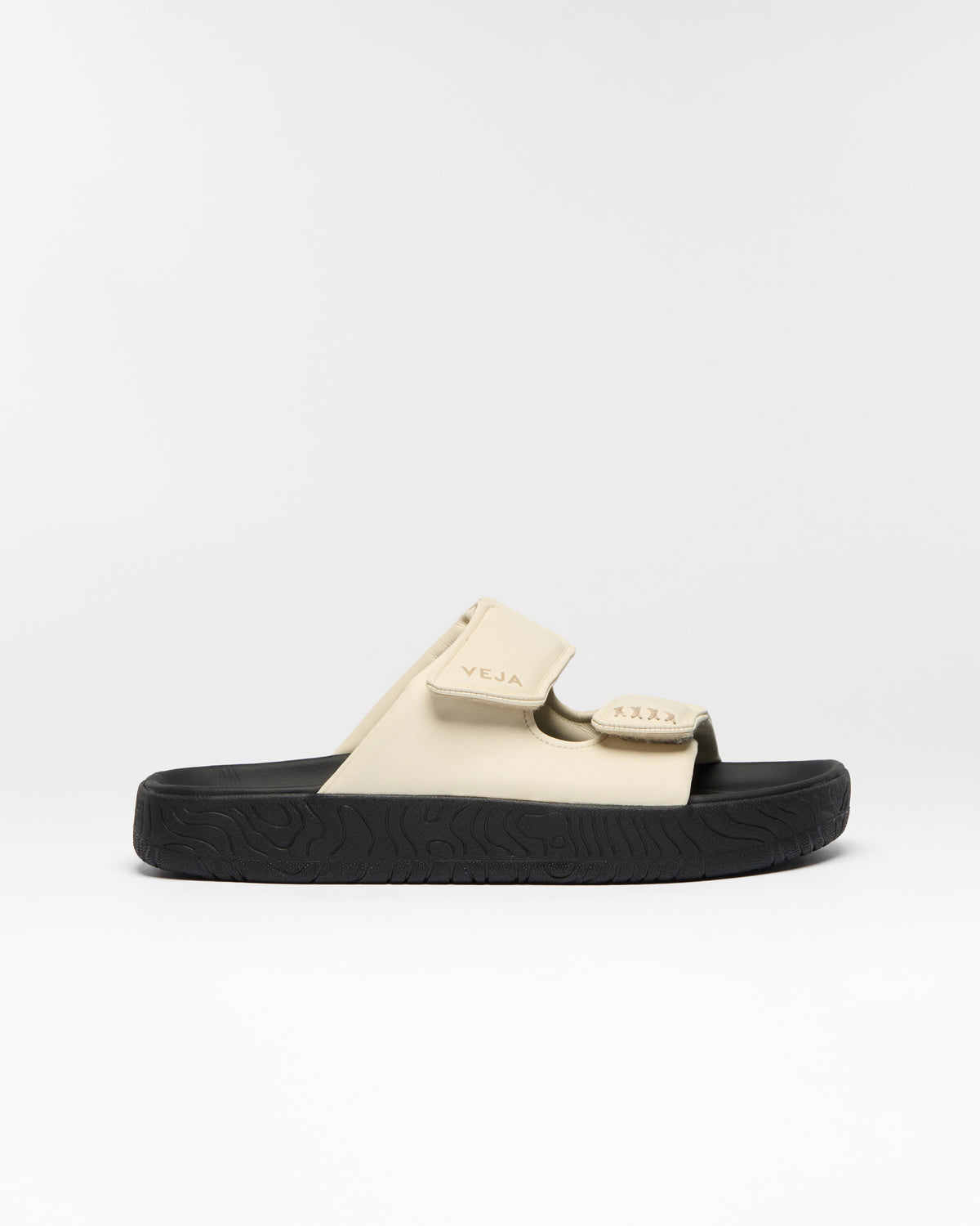 A single slide sandal by Veja, featuring a beige strap with a matching beige logo tab and a thick black textured sole, shown against a plain white studio background.