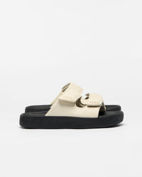 A pair of platform slide sandals with cream-colored straps and thick black soles, shown from a side profile on a light gray background.