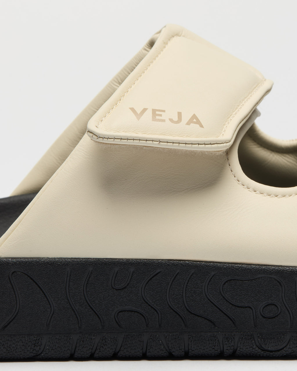 Close-up of a beige slip-on sandal with a black textured sole, showing the strap and embossed 'VEJA' branding on a light gray background.