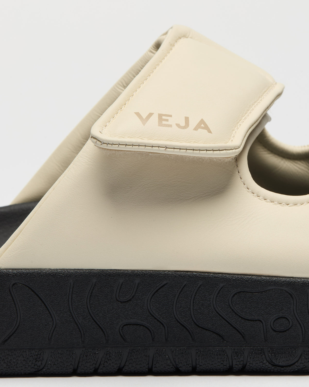 Close-up of a beige slip-on sandal with a black textured sole, showing the strap and embossed 'VEJA' branding on a light gray background.