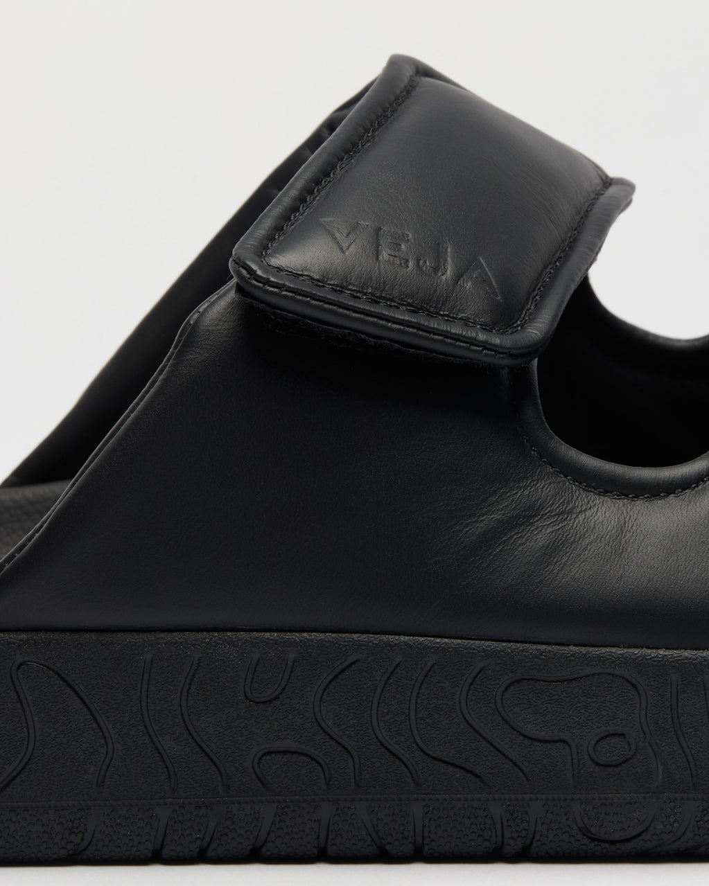 Close-up of a black leather slide sandal with a hook-and-loop strap and a thick textured black sole, featuring the brand name 'VEJA' embossed on the strap.