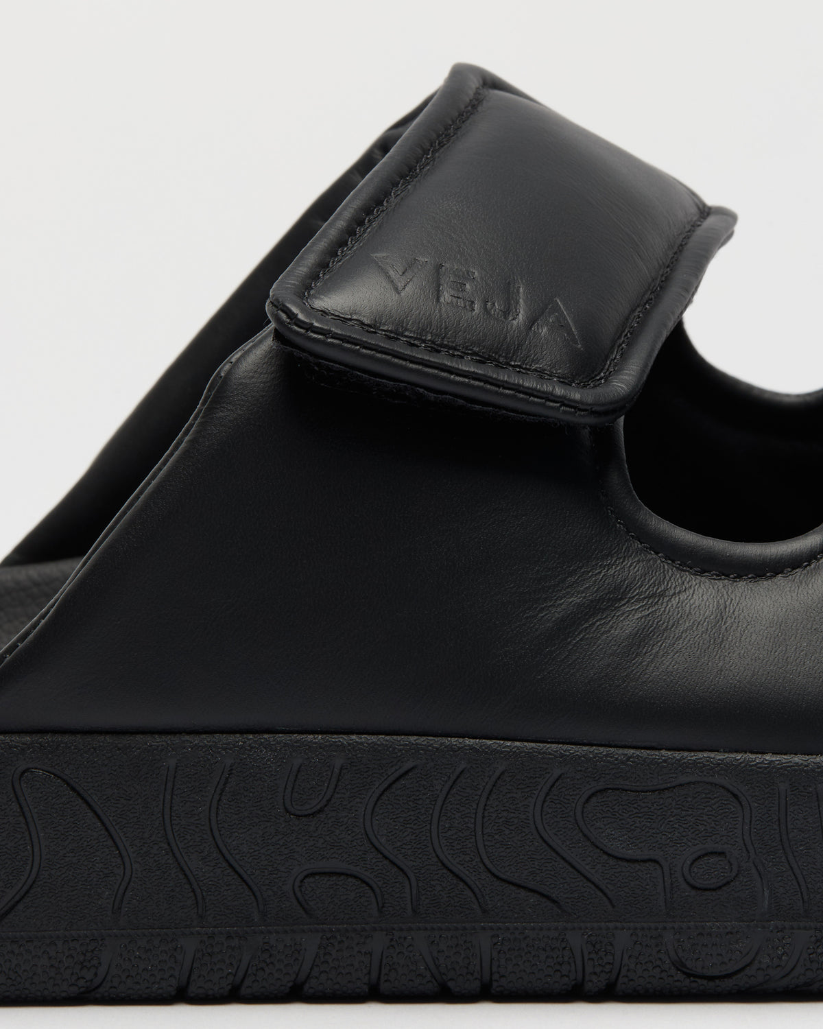 Close-up of a black leather slide sandal with a hook-and-loop strap and a thick textured black sole, featuring the brand name 'VEJA' embossed on the strap.