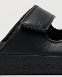 Close-up of a black leather slide sandal with a hook-and-loop strap and a thick textured black sole, featuring the brand name 'VEJA' embossed on the strap.