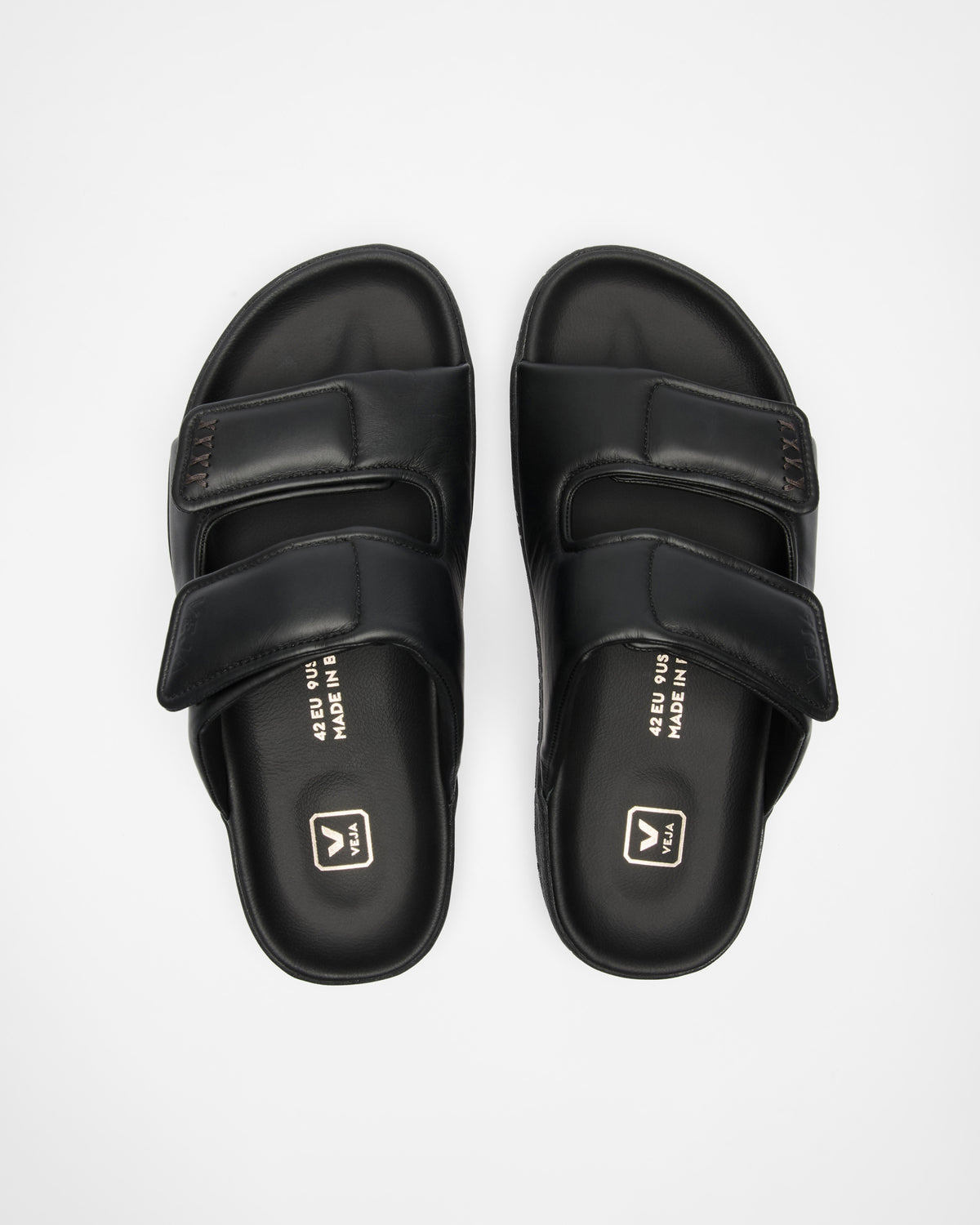 A pair of black double-strap slide sandals with a matte finish, featuring a small white logo, photographed from a top-down angle on a light gray background.