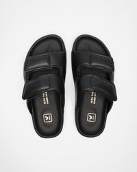 A pair of black double-strap slide sandals with a matte finish, featuring a small white logo, photographed from a top-down angle on a light gray background.