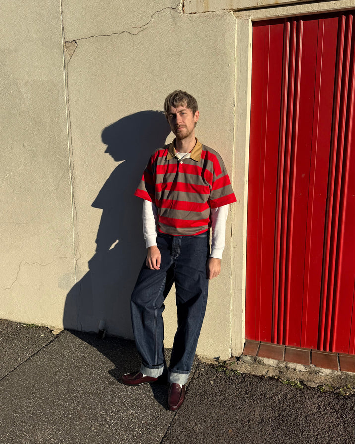 A man stands outdoors against a beige wall and a red corrugated metal door, wearing a red and gray striped short-sleeve polo shirt over a white long-sleeve crewneck, dark blue jeans, and burgundy loafers.
