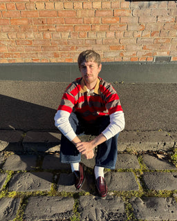 A man crouching outdoors on a stone pavement in front of a brick wall, wearing a red, brown, and beige striped polo shirt over a white long-sleeve, dark blue jeans, white socks, and burgundy loafers.