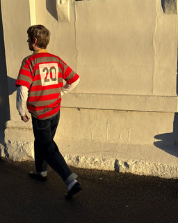 A person walking outdoors in warm sunlight wearing a red and gray striped long-sleeve jersey with the number 20 on the back, over a white long-sleeve shirt and dark jeans.