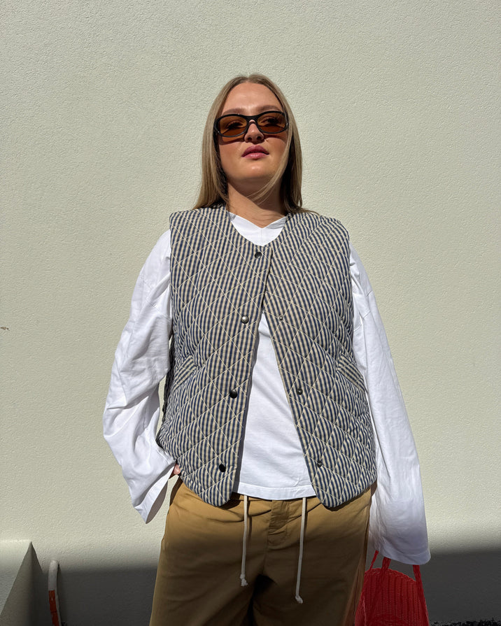 A woman stands outdoors against a plain light wall wearing a gray and white patterned vest over a white long-sleeve blouse, tan wide-leg pants, and black sunglasses. She is holding a bright red tote bag.