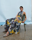 Women sits in green chair wearing a floral dress and grey suede sandals.