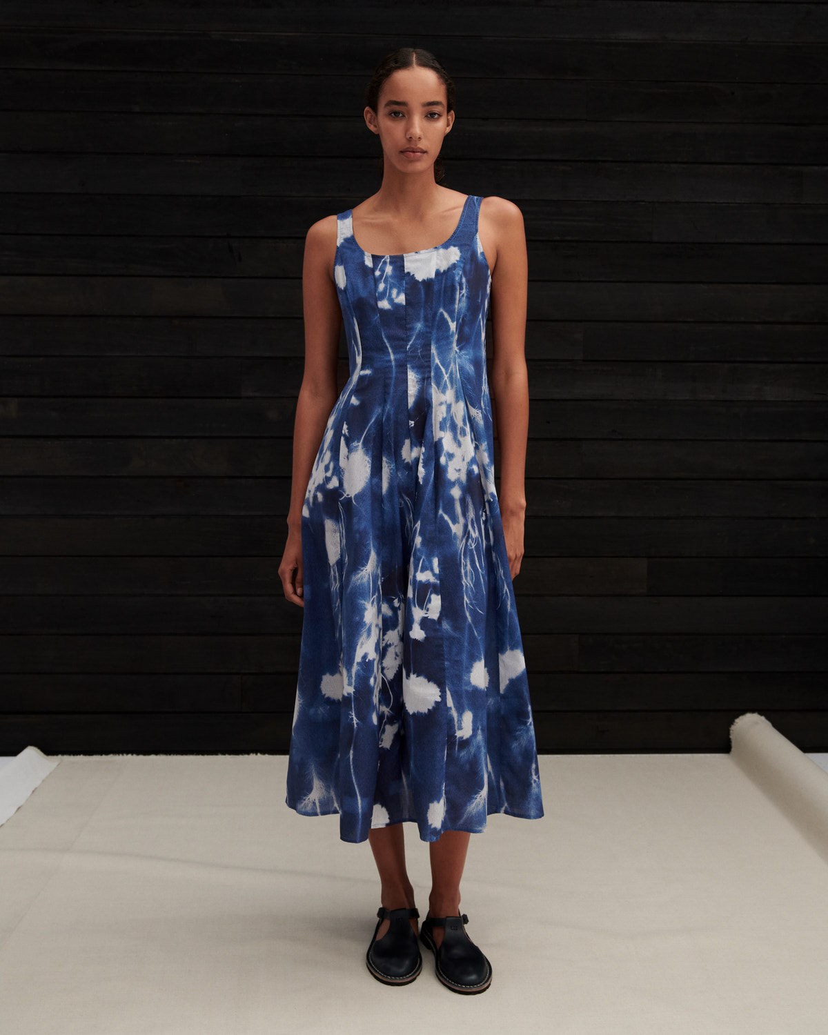 A woman modeling a sleeveless midi dress with a blue and white tie-dye pattern, standing against a dark wood-paneled wall.