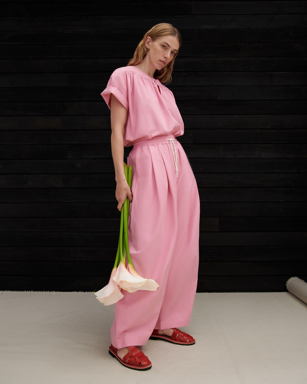 A woman modeling a light pink two-piece linen set consisting of a short-sleeve top and matching wide-leg trousers, holding a bunch of white calla lilies against a dark wood-paneled background.
