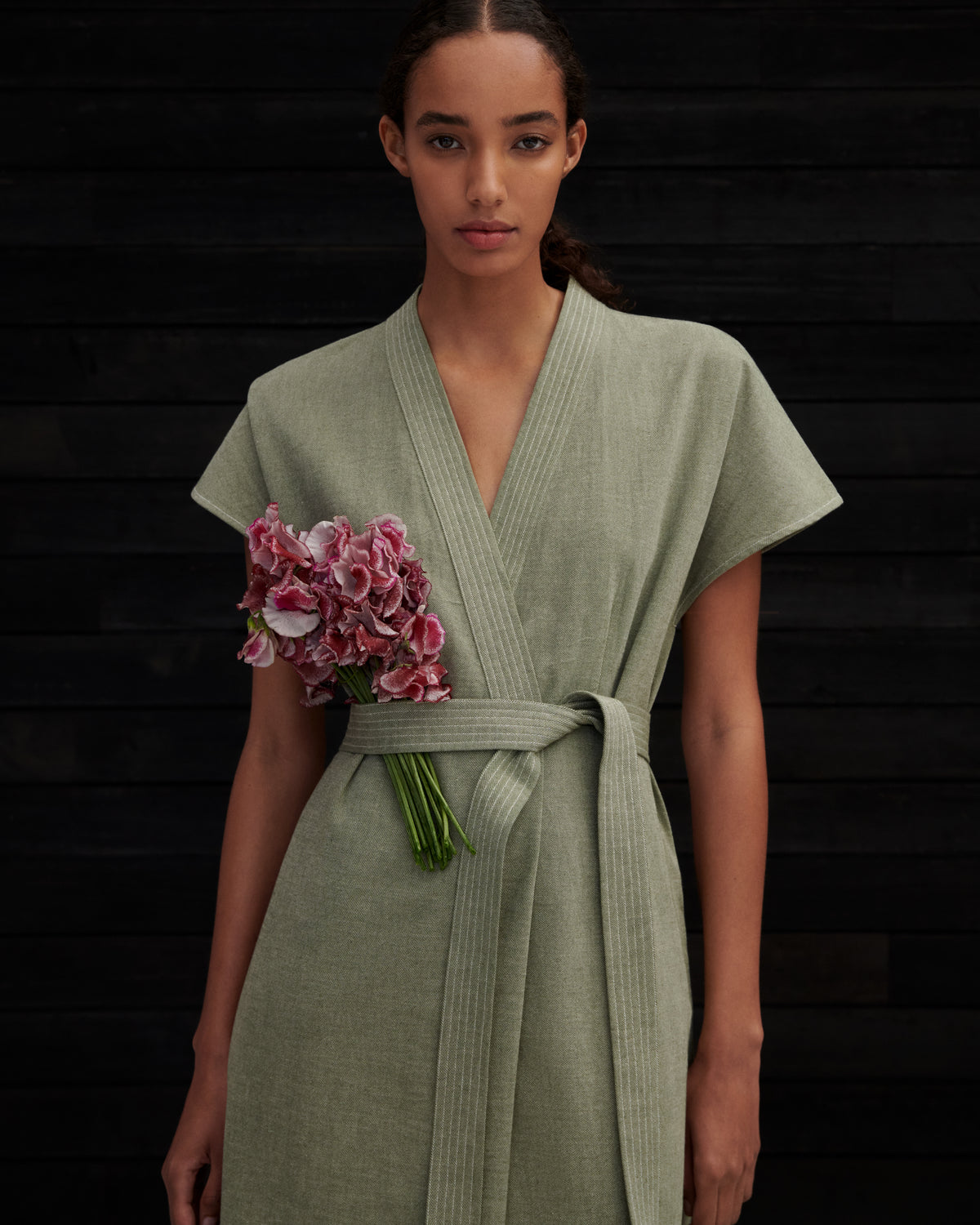 A woman wearing a sage green linen wrap robe with a matching tie belt, holding a bouquet of pink flowers against a dark wood plank background.