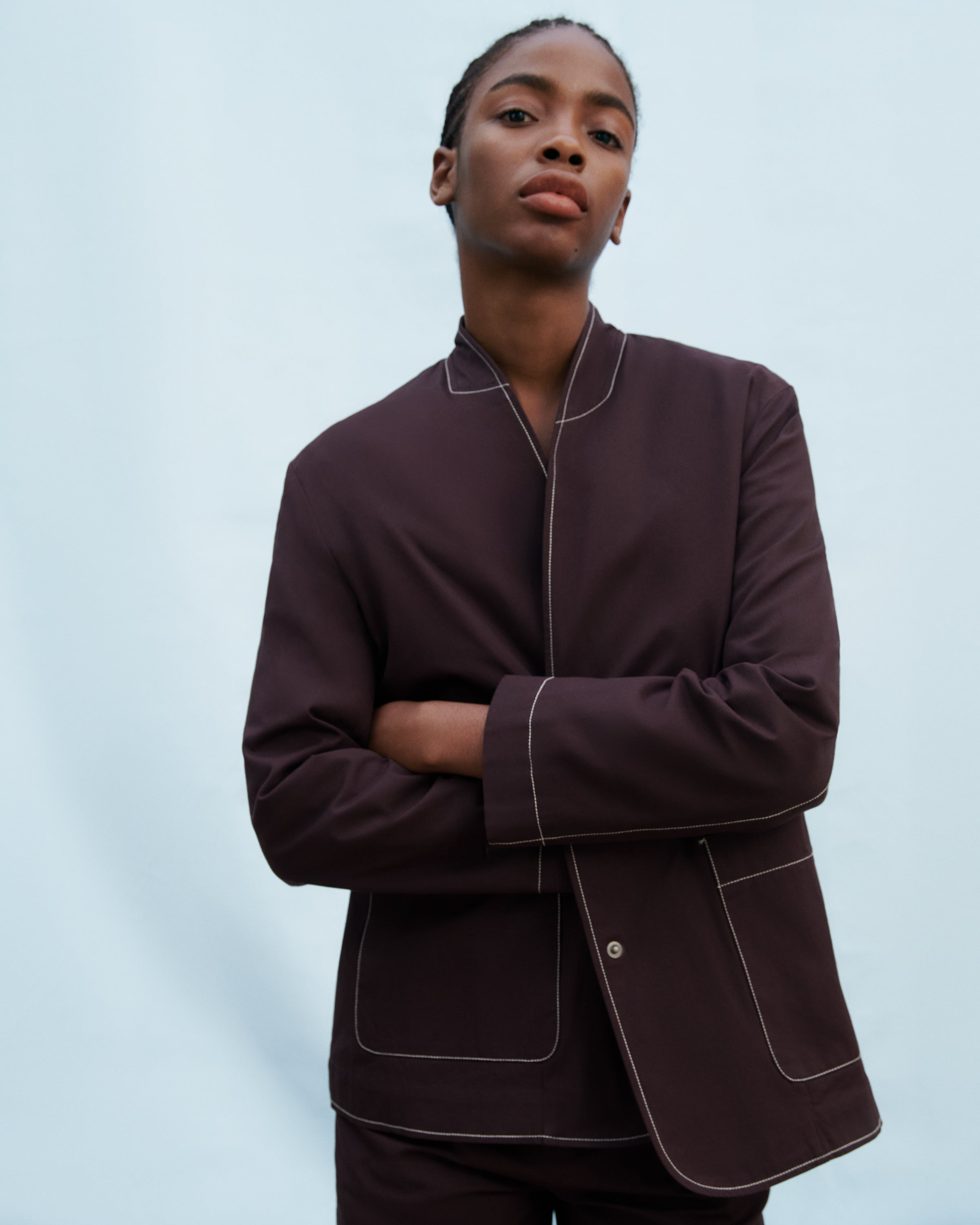 A model wearing a dark plum-colored pajama-style jacket with contrasting light piping, posed against a solid light blue background.