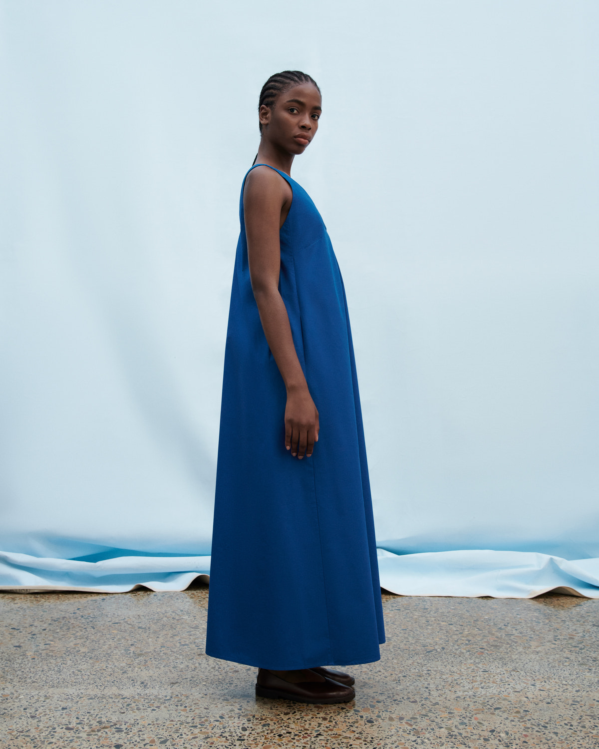 A model wearing a sleeveless, floor-length blue dress stands in profile against a light blue backdrop with a draped fabric floor.