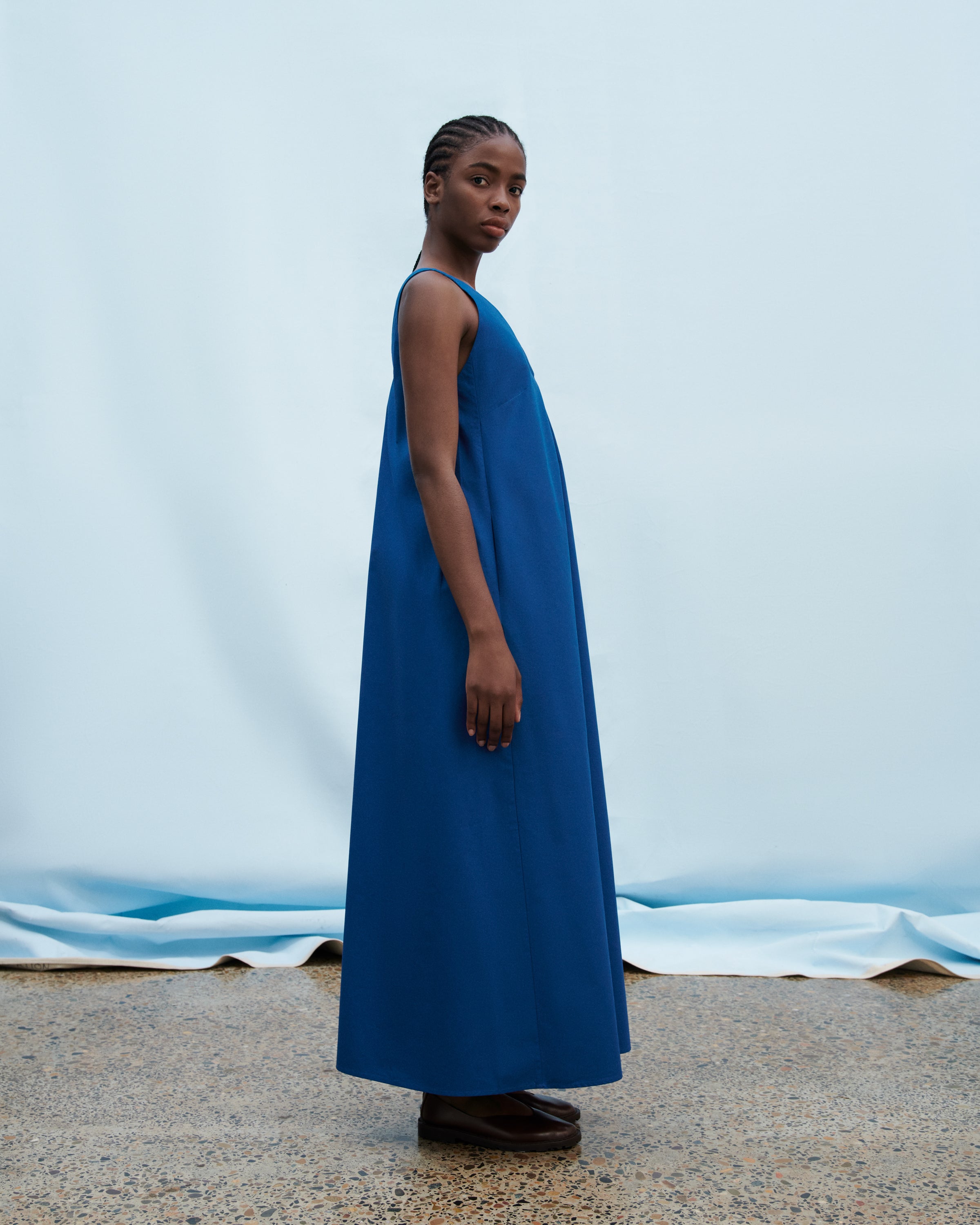 A model wearing a sleeveless, floor-length blue dress stands in profile against a light blue backdrop with a draped fabric floor.