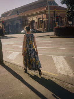 A woman wearing a sleeveless blue and yellow patterned maxi dress walks across a city crosswalk in front of a brick building.