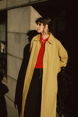A woman wearing a long tan trench coat over a bright red top and black pants, standing outdoors against a stone building wall in dramatic sunlight and shadow.