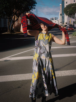 A woman stands on a city street crosswalk wearing a sleeveless, high-neck maxi dress with a gray, yellow, and white abstract pattern. She is holding up a red and black patterned knit cardigan or sweater over her head in a playful pose.