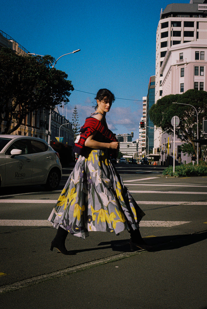 A woman walks across a city street crosswalk wearing a red top and a long blue dress with yellow, white, and purple iris's. The background features urban buildings, trees, and a clear blue sky.