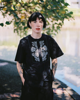 A person with short dark hair stands outdoors under leafy trees near a body of water, wearing a loose black dress with white floral embroidery around the neckline and on the lower front.