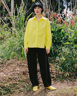 A person standing outdoors in a garden wearing a bright yellow button-up shirt, black wide-leg pants, and matching yellow shoes.