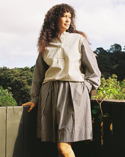 A woman with long curly hair stands outdoors wearing a matching striped button-front blouse and pleated skirt set. The outfit features vertical black and off-white stripes, set against a background of trees and a cloudy sky.