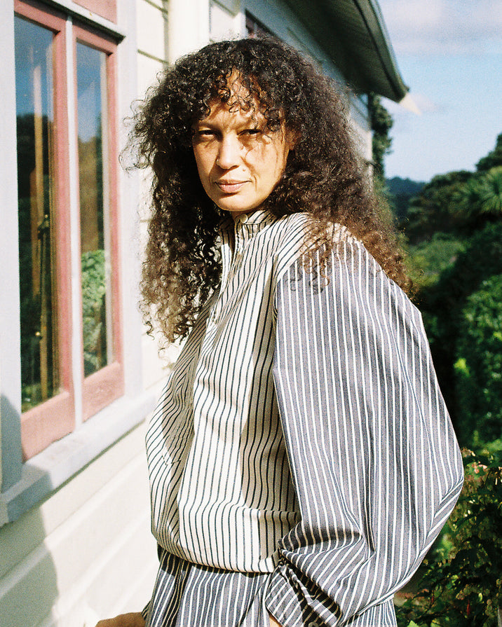 A woman with long, curly dark hair stands outdoors in front of a house, wearing a loose, high-neck striped top and matching bottoms. The outfit features vertical stripes in gray and off-white.
