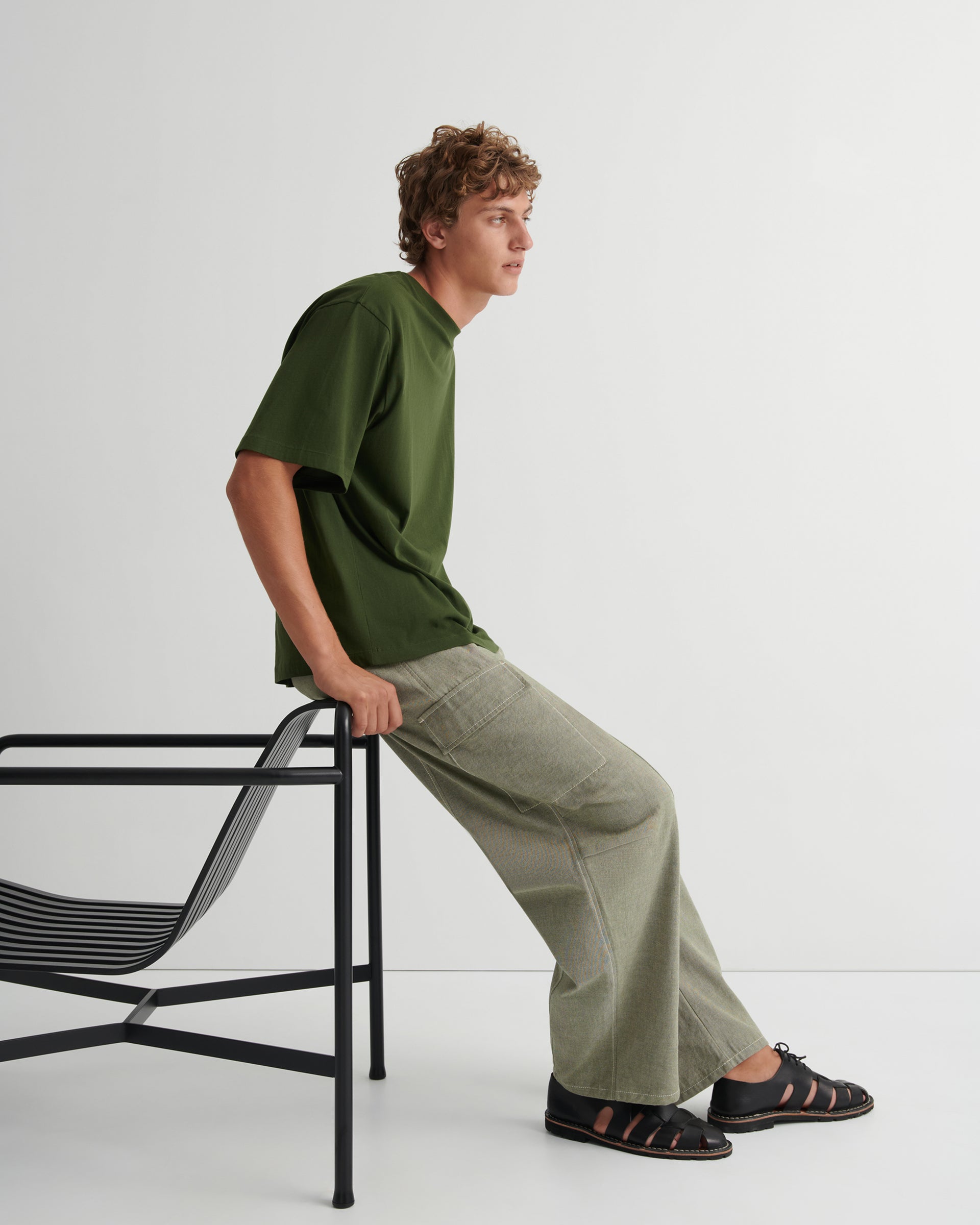 Man wearing a dark green oversized tee and sage green trousers with black sandals on, leans on a black deckchair against a grey background.