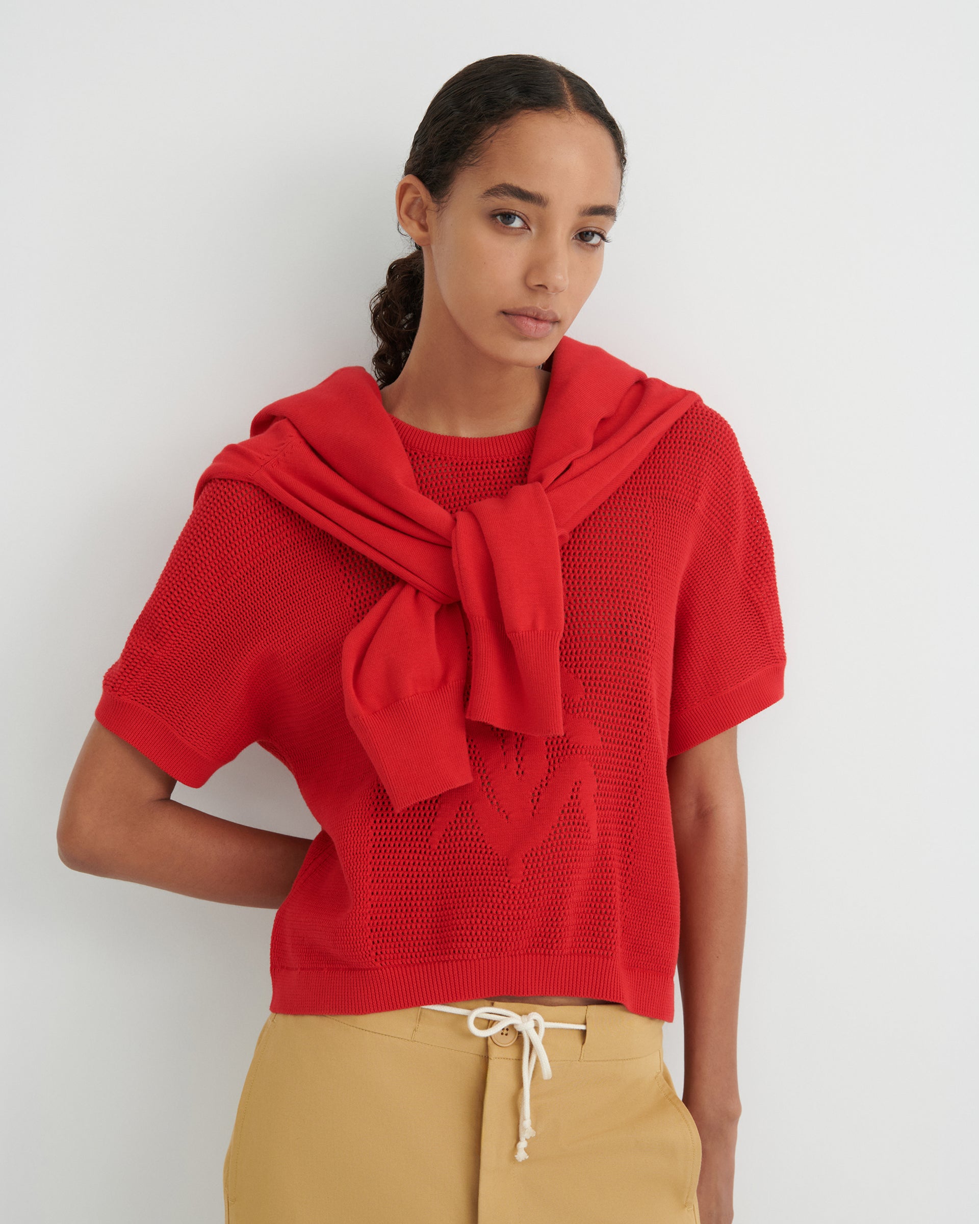 Women wearing a red knit tee and beige trousers with a white drawstring and a red sweater draped on her shoulders.