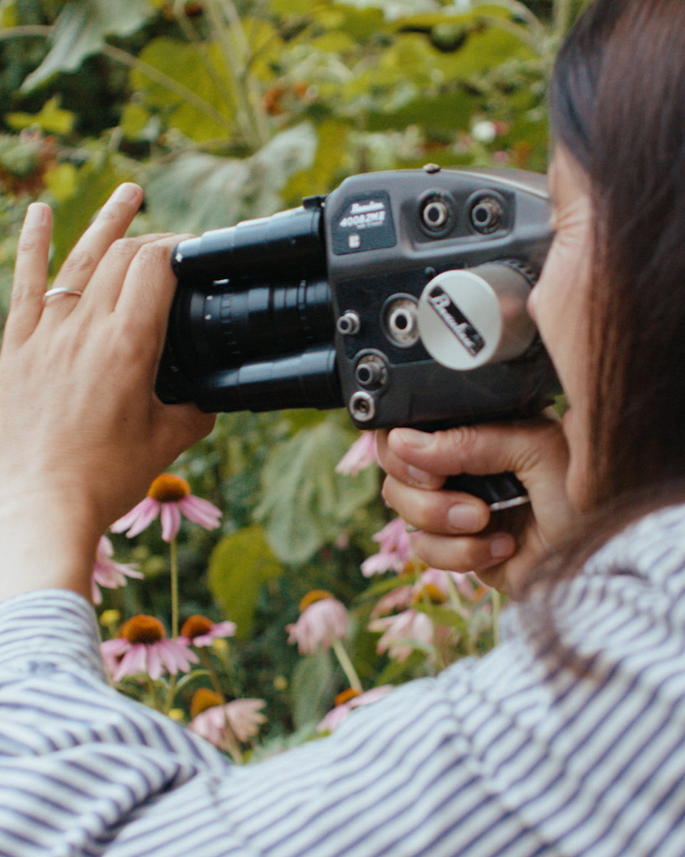 A person in a striped shirt holding a black and gray video camera up to their eye, photographed from behind, with green foliage and pink coneflowers in the background.