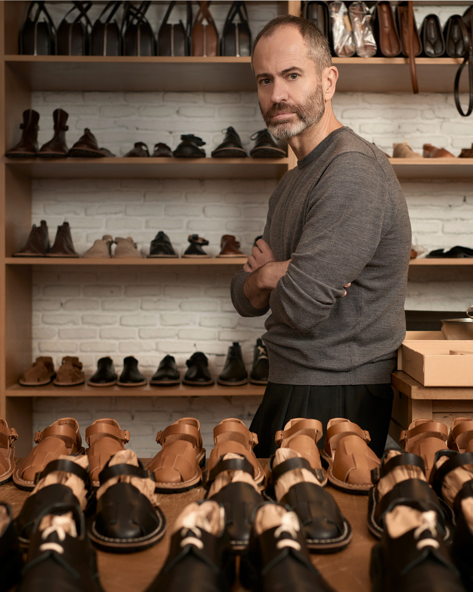 A man with a graying beard and short hair stands in a shoe store, wearing a gray long-sleeve shirt and black trousers. Shelves behind him display various pairs of shoes, and several tan and black leather shoes are arranged in the foreground